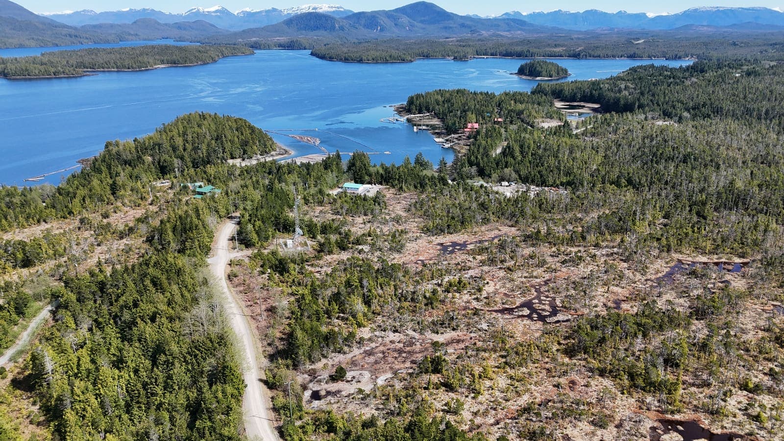 Aerial view of a west coast Vancouver Island inlet with forested islands, a small marina and snow-capped mountains on the horizon