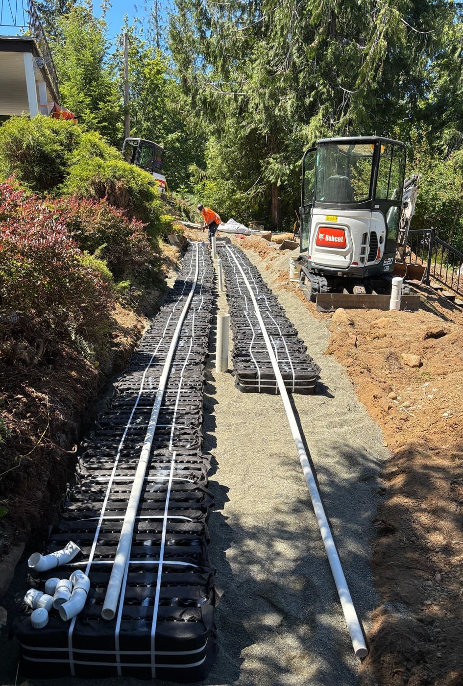 Rows of black proprietary sand-filter dispersal modules laid in a narrow bed on a sloped property, with white PVC distribution pipe running the length and a compact excavator working alongside