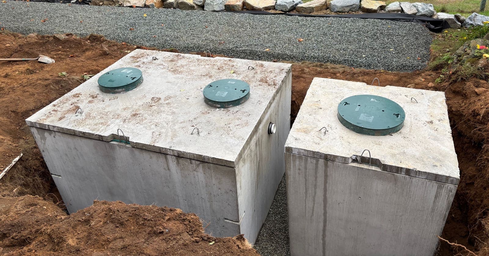 Two concrete septic tanks with green access lids set side by side in a fresh excavation, backfill gravel and a rock retaining wall behind them