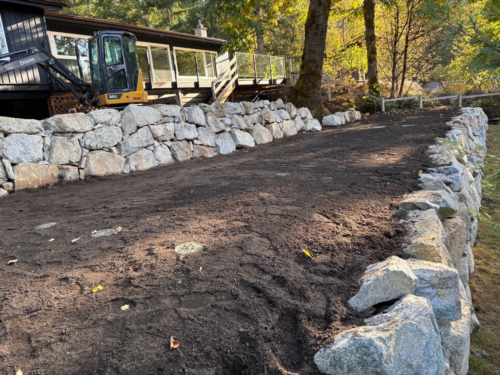 Completed terraced dispersal bench on a sloping property, bounded by two courses of granite armour rock retaining wall, with graded soil cover and observation port lids visible at the surface and a compact excavator beside the house