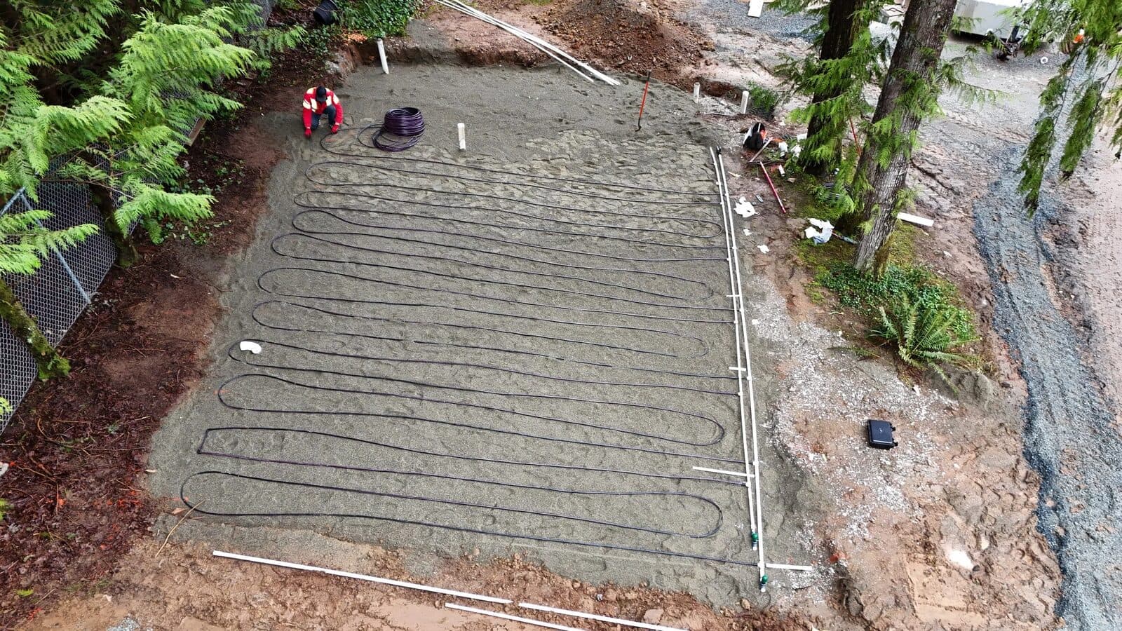 Aerial view of a rectangular dispersal bed of levelled sand with black drip tubing laid in long loops, bounded by white PVC pipe and surrounded by forest