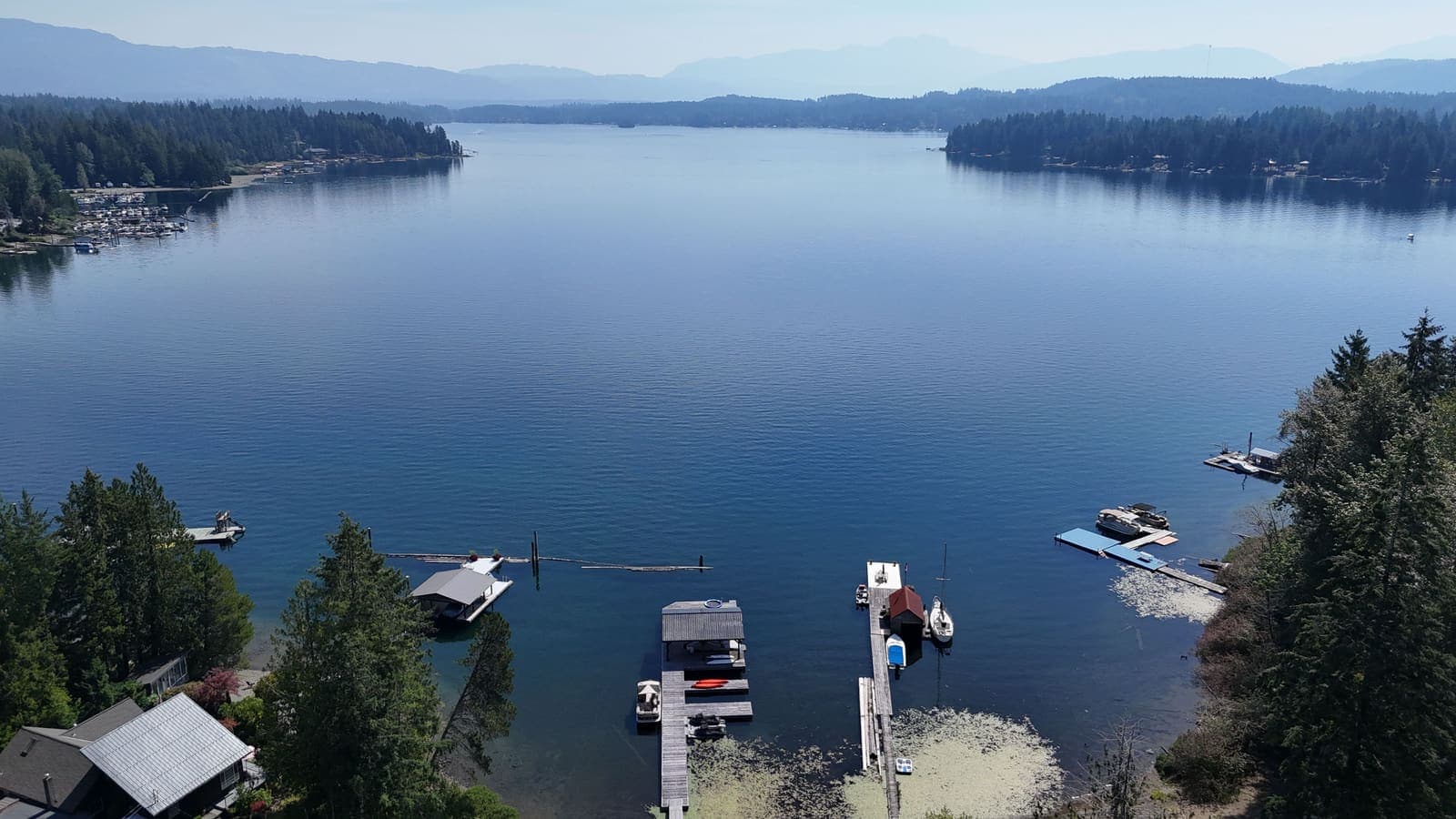 Aerial view of a large Vancouver Island lake ringed by forest and mountains, with private docks along the near shoreline