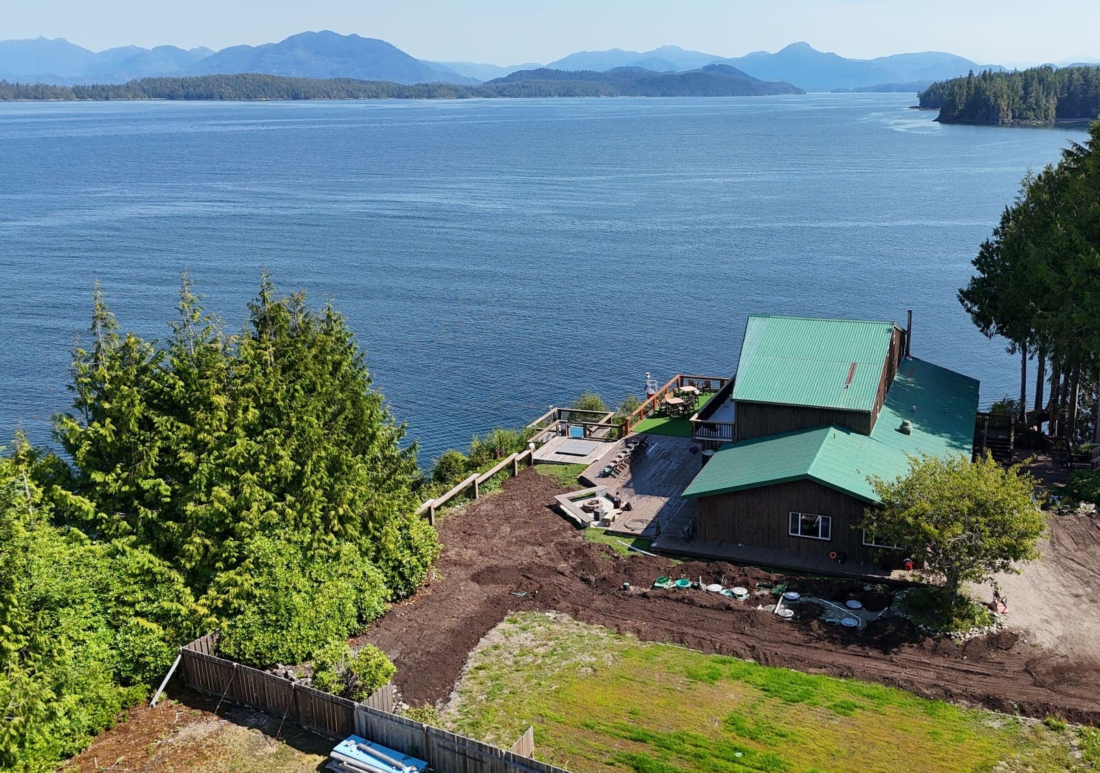 Aerial view of a remote west coast waterfront lodge with a green metal roof, freshly excavated ground for a septic installation in the foreground and forested islands across a calm inlet