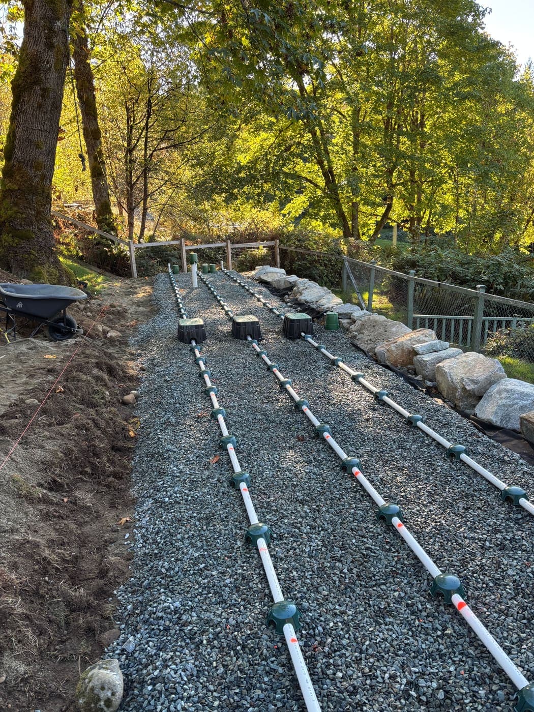 Three long white PVC pressure distribution laterals with green orifice shields running the length of a gravel dispersal bed on a wooded slope