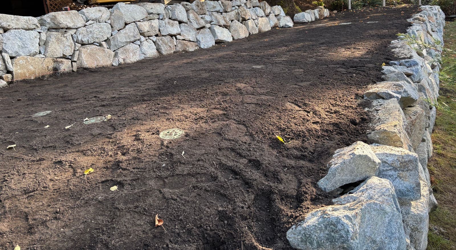 A completed dispersal field covered with dark topsoil and graded flat between two granite rock walls, with observation port lids flush at the surface