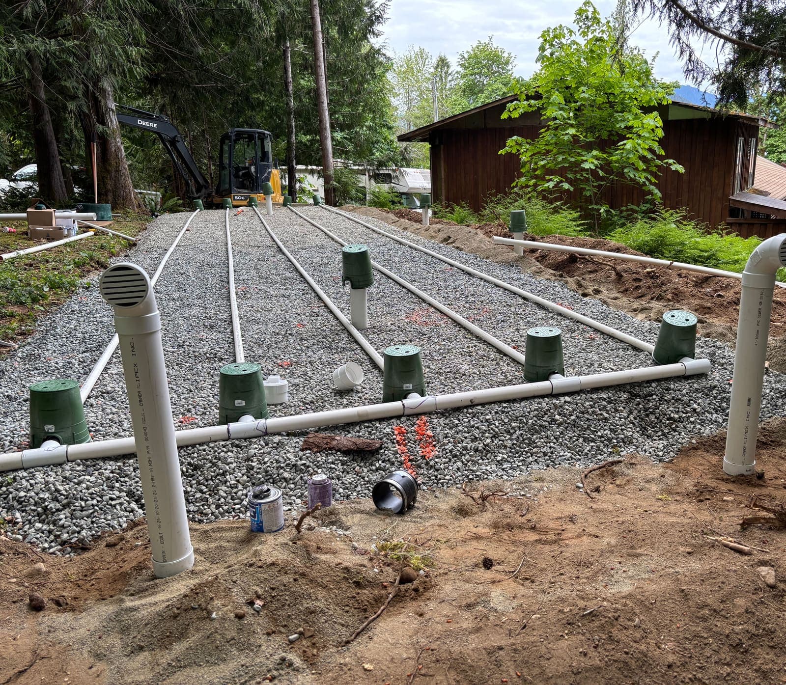 Sand lined seepage bed under construction with white PVC pressure laterals, green observation port risers and two tall passive vent risers, an excavator parked at the head of the bed