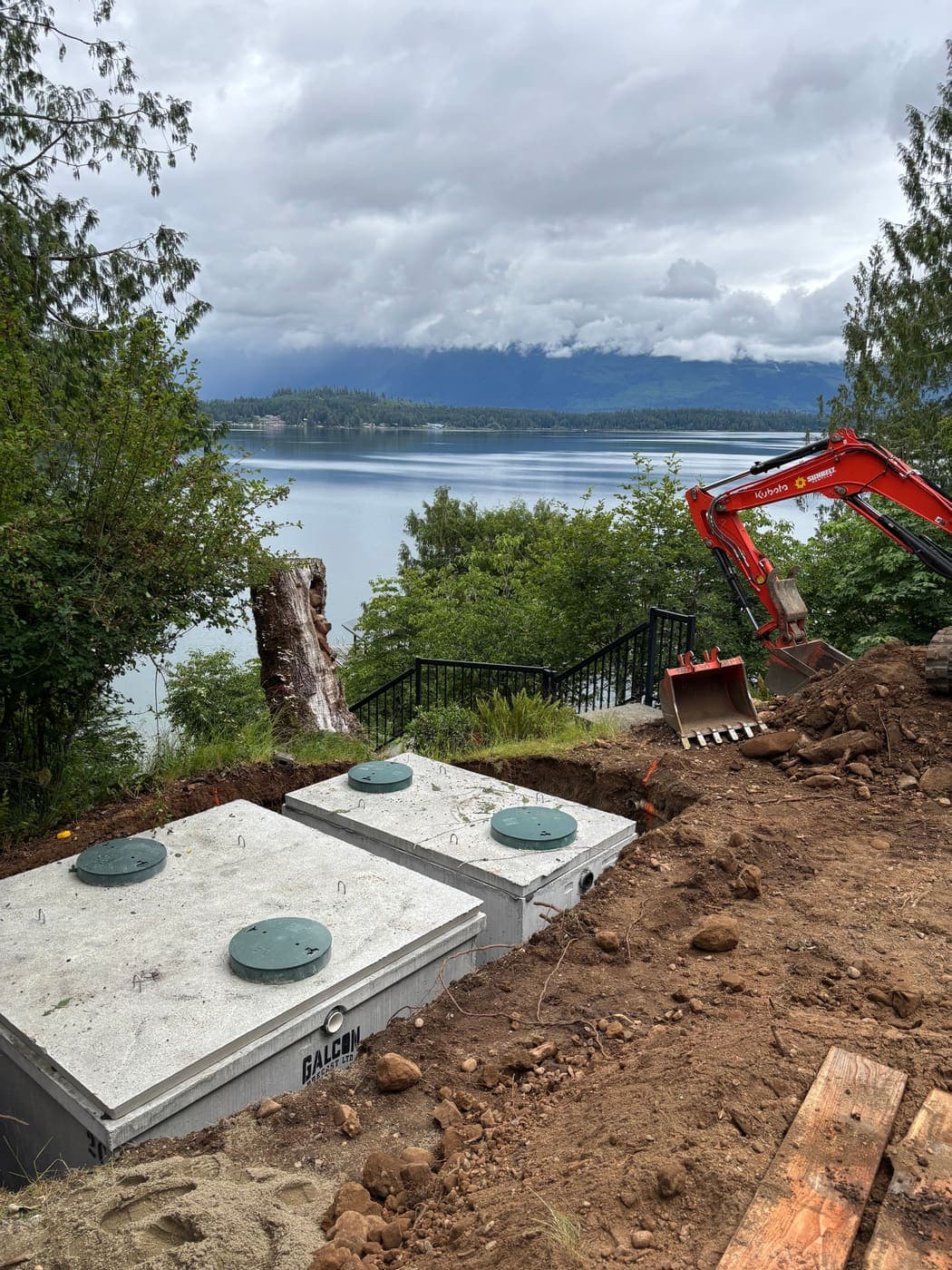 Two concrete septic tanks with green riser lids set into an excavation on a lakeside bench, with a red excavator alongside and forested mountains across the water