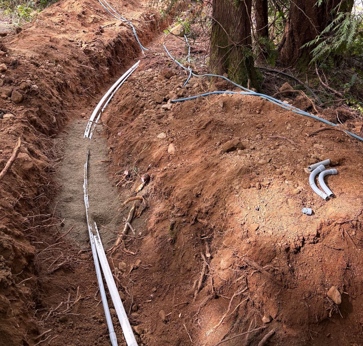 An open excavated trench on a forested slope with white PVC supply lines bedded in imported sand and grey electrical conduit running along the bank above
