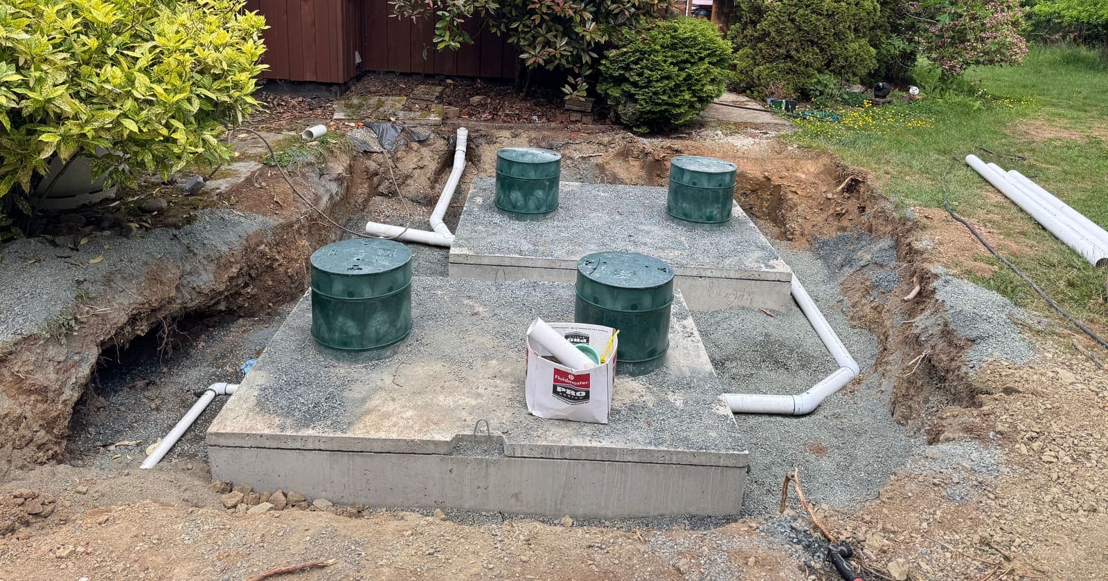Concrete tank lids with four green access risers set on a levelled gravel pad in an excavation beside a house, with white PVC pipework running to and from the tanks