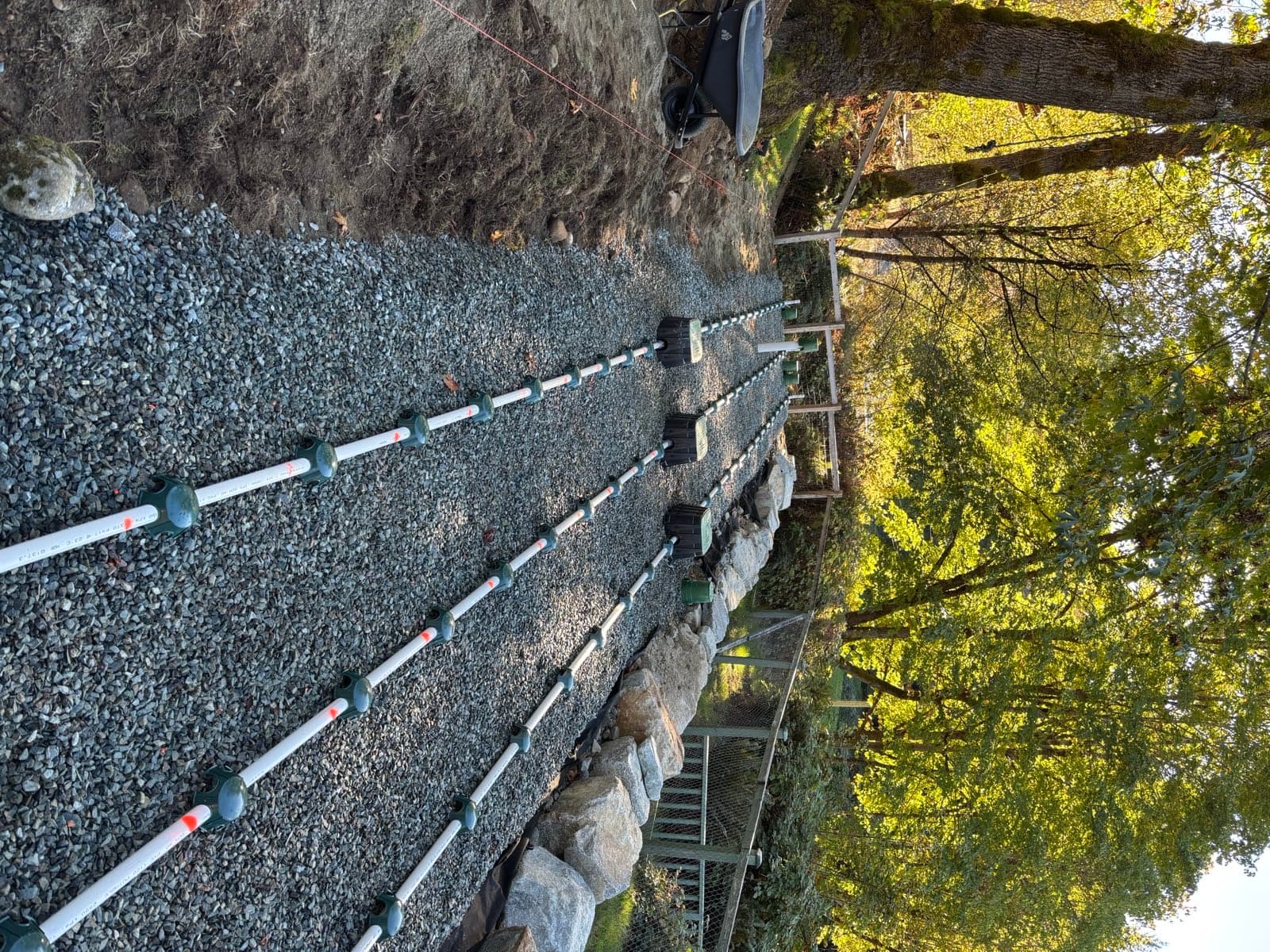 Three white PVC pressure distribution laterals with orifice shields running the length of a washed drain rock bed on a terrace, with valve boxes at the head and a granite rock retaining wall along one edge