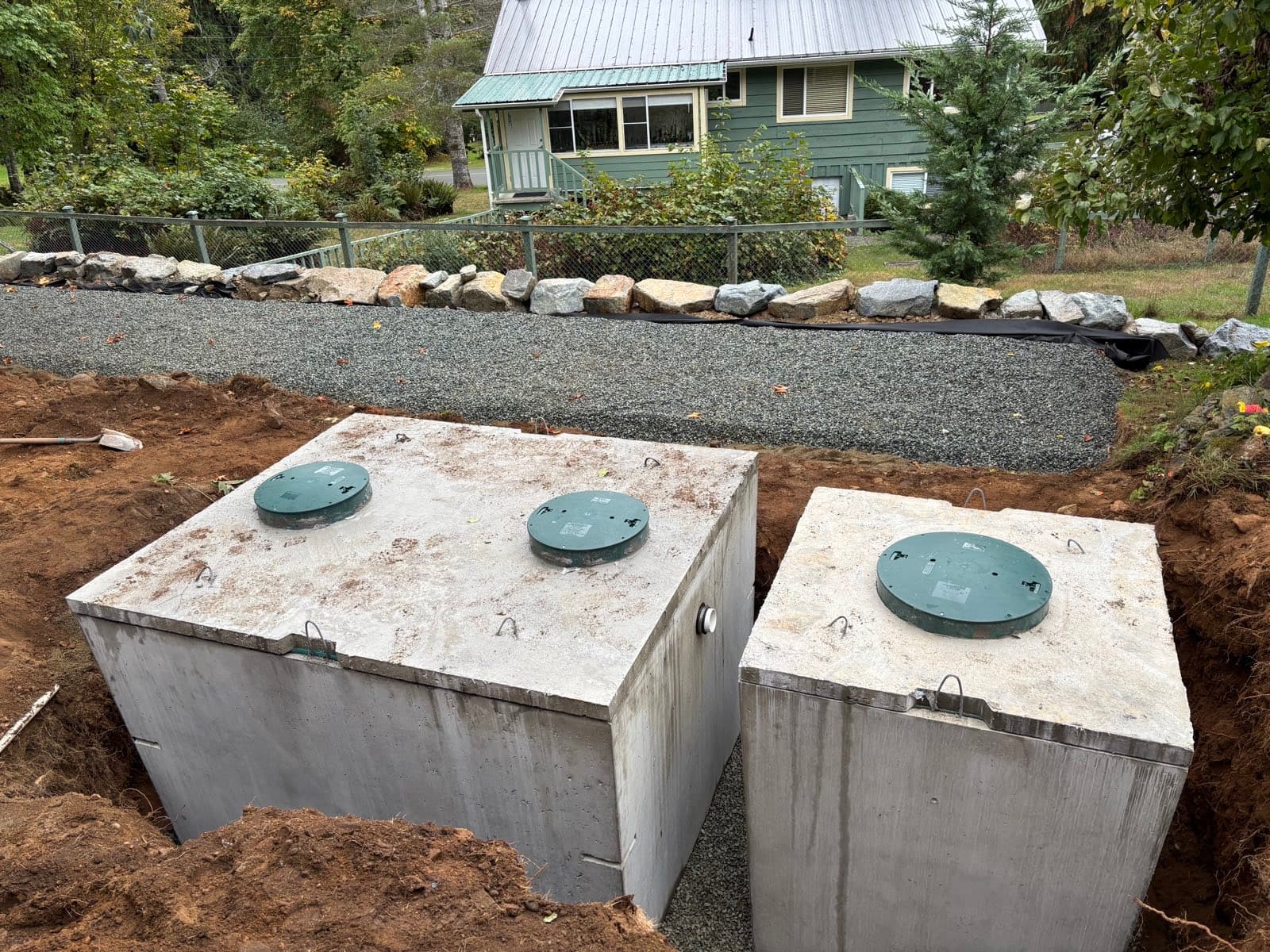 Two large concrete septic tanks with green access lids set side by side in an excavation below a rock retaining wall and gravel terrace, with a house behind