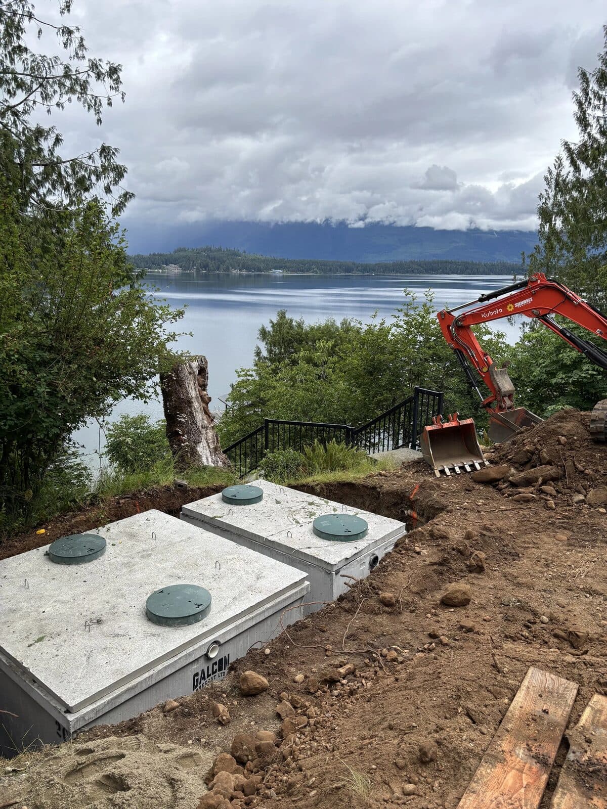 Two newly set concrete septic tanks in an open excavation above a Vancouver Island lakeshore, with an excavator working alongside