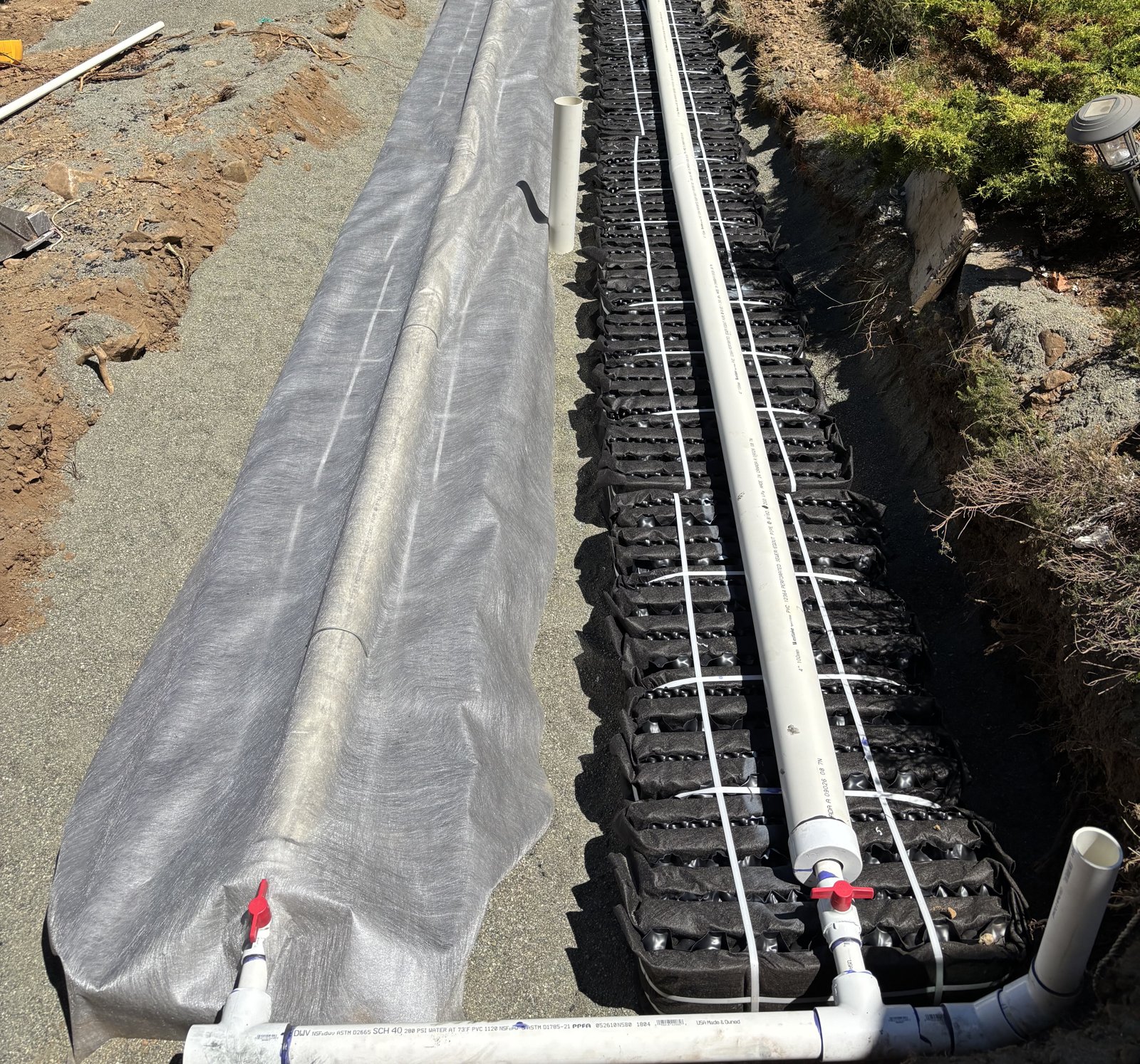 Two rows of combined treatment and dispersal modules in a sand bed, one row wrapped in geotextile, with a white pressure distribution pipe running along the top of each row and valved flushing ends in the foreground