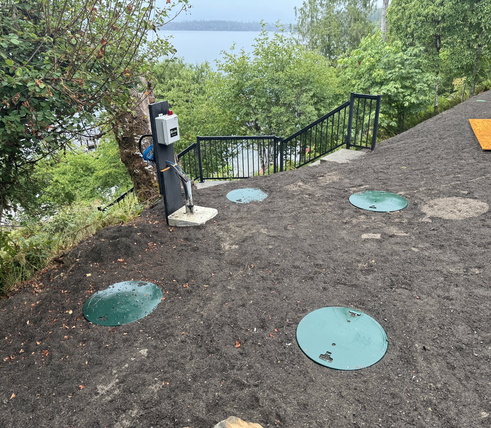 Finished lakefront septic site on a bench above a lake, with green tank lids set in fresh soil, a control panel on a post, and a railing and stairs leading down toward the water through the trees